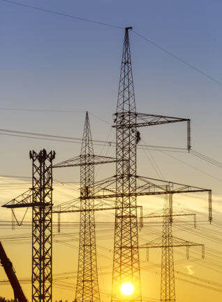 Stromleitungen und Freileitungsmasten im Sonnenuntergang mit Monteuren bei Arbeiten an der Leitung.