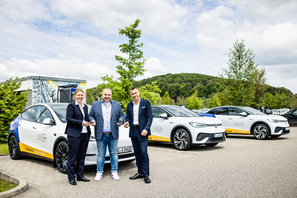 Cteam: trois personnes posant devant des voitures électriques sur un parking, avec une borne de recharge en arrière-plan.